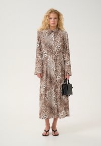 Abito in stampa leopardata con maniche lunghe, colletto e cintura in vita. La modella tiene una borsa nera e indossa sandali neri. Tessuto dalla texture liscia.