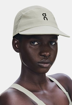Personne à la peau foncée portant une casquette beige clair avec un logo à l'avant et des panneaux latéraux perforés, ainsi que des bretelles de débardeur beige clair assorties.