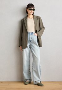 Blazer verde oliva, blusa beige, jeans de tiro alto en azul claro y zapatos tipo slip-on verdes. Los accesorios incluyen gafas de sol oscuras y pendientes.