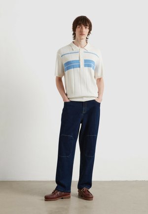 Junger Mann steht mit den Händen in den Taschen, trägt ein cremefarbenes, gestricktes Polo mit blauen Streifen, dunkle Jeans mit Knieflicken und braune Lederschuhe.