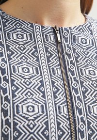 Granatowa tkanina z białymi geometrycznymi wzorami, z metalowym zamkiem błyskawicznym z przodu. Tekstura wydaje się tkana, z wyraźnymi detalami.