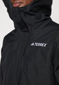 Sort Terrex jakke med høj krave, justerbar hætte og broderet logo. Materialet er glat med mat finish. Skjult lynlåslomme.