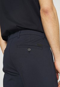 Pantalon bleu marine en tissu texturé, avec une poche arrière dotée d'un bouton noir et d'une petite étiquette de marque. Conception ajustée.