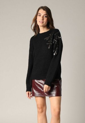 Pull en tricot noir avec une texture duveteuse, arborant un motif de feuille en sequins sur une épaule. Associé à une mini-jupe bordeaux brillante.