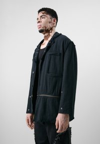 Schwarze Jacke mit ausgefransten Kanten, Reißverschluss, Druckknöpfen und zwei vorderen Taschen. Getragen über einem schwarzen Tanktop und zerrissenen schwarzen Hosen.
