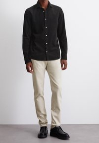 Camisa negra de botones con mangas largas, combinada con pantalones ajustados beige claro y zapatos negros, en un fondo neutro.