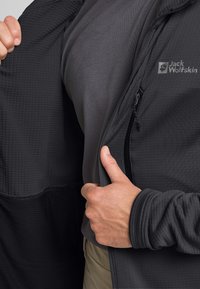 Veste noire en tissu texturé, dotée d'une fermeture éclair et d'un logo. En dessous, une chemise sombre lisse est partiellement visible.