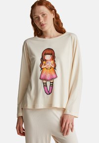 Eine rothaarige Frau trägt ein cremefarbenes Langarmshirt mit einem Grafikdruck, der ein gesichtsloses Mädchen in einem orange-pinken Kleid zeigt, das ein Glas hält.