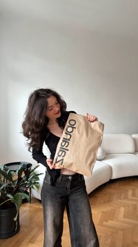 Junge Frau mit dunklen Haaren hält eine große braune Papiertüte mit schwarzem Text und steht in einem Wohnzimmer mit einem weißen Sofa und einer Pflanze.