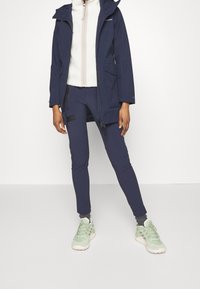 Marinkapad jacka med huva, kombinerad med marinblå tajta byxor och ljusgröna sneakers. Har en texturerad krämfärgad inredning.