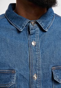 Blå denimskjorte med spiss krage, metalliske trykknapper og doble brystlommer med synlige sømdetaljer.