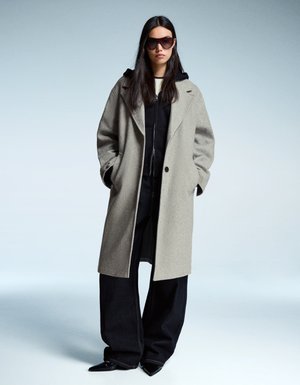 Femme portant un manteau gris clair oversize, un hoodie noir, un pantalon noir à larges jambes, des chaussures à bout pointu et de grandes lunettes de soleil sombres sur un fond clair.