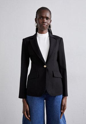 Black tailored blazer with a single gold button, notched lapels, and two front pockets, paired with a white high-neck top and blue jeans.