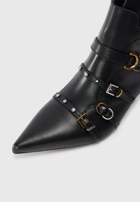 Botín de cuero negro hasta el tobillo con punta afilada, múltiples correas, hebillas de oro y plata, y acentos de tachuelas. Textura suave, diseño elegante.