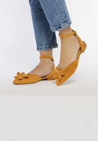 Bailarinas amarillas de ante con diseño floral en la puntera, forma puntiaguda, tira en el tobillo y tacón bajo. Combinadas con unos vaqueros azules remangados.