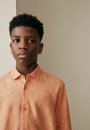 Lühikeste lokkis juustega poiss, kes kannab virsikuvärvi nööpidega särki ja seisab neutraalse beežika tausta ees, vaadates kõrvale.