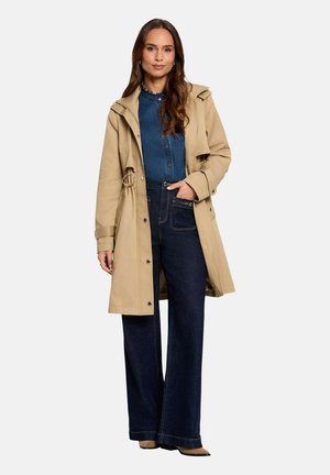 Femme portant un trench-coat beige par-dessus une chemise bleue boutonnée et un jean large sombre, debout avec une main dans la poche.