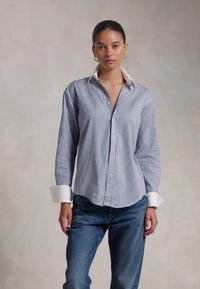 Cămașă cu dungi albastre și albe, cu guler clasic și manșete albe în contrast, purtată cu blugi de denim bleu închis.