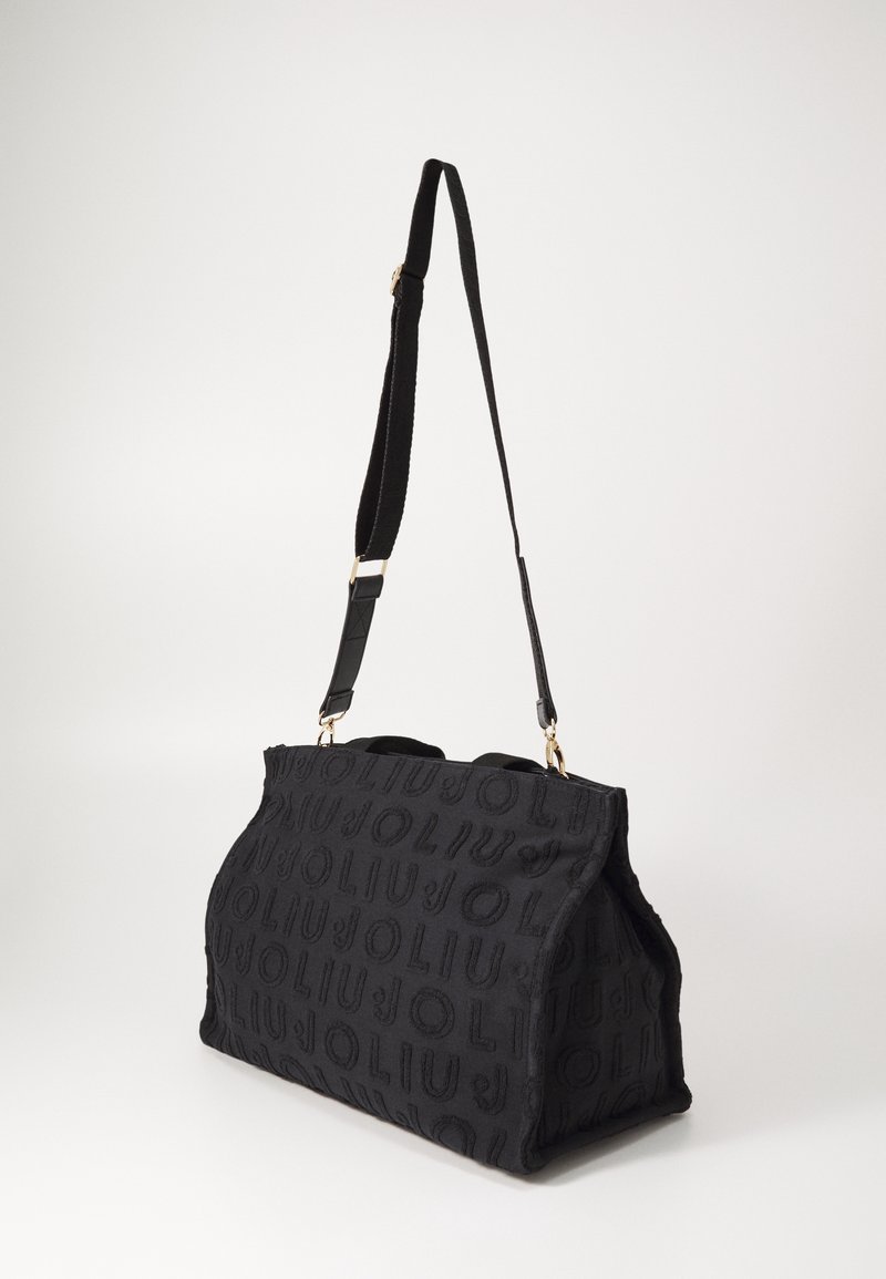 Sac d'épaule en tissu noir avec une large sangle réglable et un motif de texte embossé répétitif, présenté sur un fond clair uni.