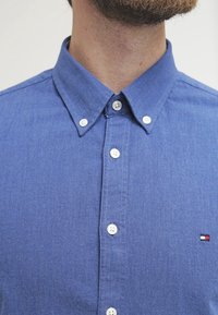 Camicia azzurra con bottoni realizzata in tessuto intrecciato. Presenta un colletto button-down, bottoni bianchi e un piccolo logo sul lato sinistro del petto.