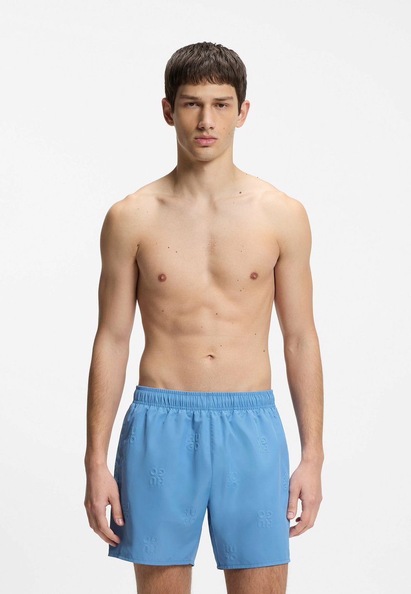 Jeune homme aux cheveux courts et foncés, debout face à l’avant, torse nu, portant un short de bain bleu clair avec texte embossé sur fond blanc.