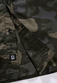 Maskáčová košeľa s tmavozelenými a čiernymi vzormi, čiernymi gombíkmi a malou čiernou štítkom s bielym logom pri spodnom okraji.