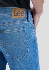 Die Hand einer Person ruht in der Gesäßtasche einer blauen Lee-Jeans mit sichtbarem Lederlogo über der Tasche.