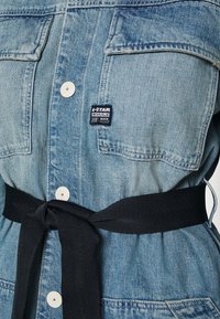 Jeansklänning i ljusblått med två bröstfickor, vita knappar och ett svart tygbälte som är knutet i midjan. Har G-Star-märke.