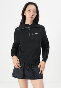 Woman wearing black zip-up Tommy Hilfiger sweatshirt and black denim skirt with large front pockets, standing against white background.