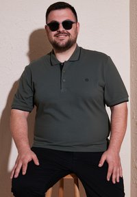 Olivovo zelená polo košeľa s triom gombíkov, krátkymi rukávmi a čiernym lemovaním. Čierny znak na hrudi. Model sediaci na drevenej stoličke.