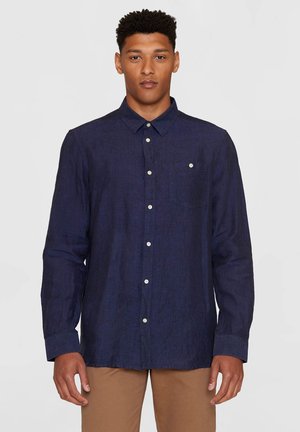 Man wearing a buttoned navy blue long-sleeve shirt with a chest pocket and tan pants, standing against a plain light background.