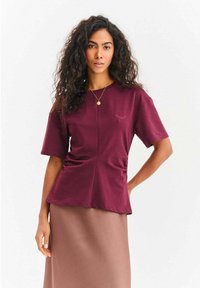 Bordowa bluzka z krótkim rękawem i okrągłym dekoltem, z marszczeniami po bokach oraz subtelnym motywem rzęs na piersi.