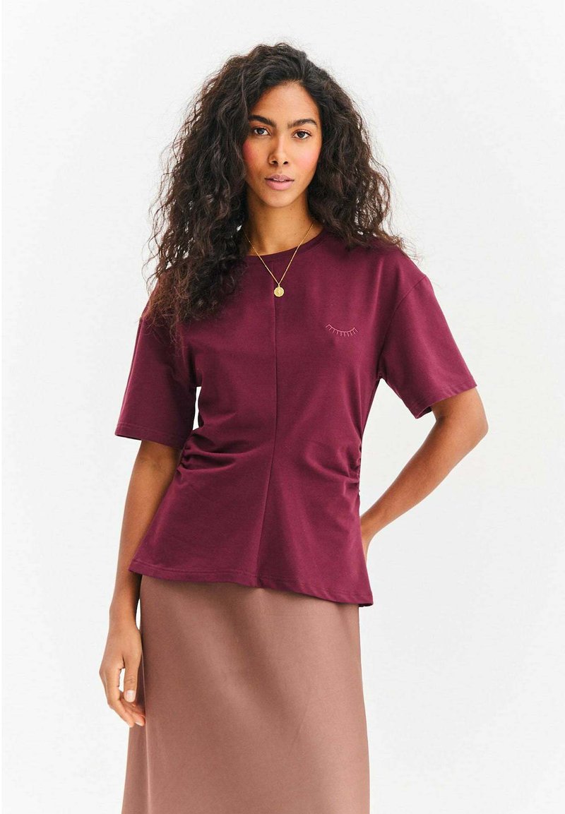 Bordowa bluzka z krótkim rękawem i okrągłym dekoltem, z marszczeniami po bokach oraz subtelnym motywem rzęs na piersi.