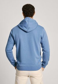 Muž s kudrnatými vlasy, oblečený v světle modré mikině a béžových kalhotách, stojící čelem k odvrácené straně s rukama v kapsách na pozadí bez vzoru.