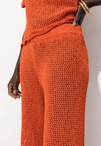 Ensemble en maille orange comprenant un haut et un bas, avec un design texturé et en tricot aéré. Le haut a un col rond, et le pantalon est à coupe décontractée.