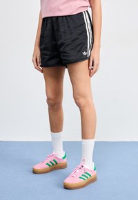 Black athletic shorts with white side stripes and Adidas logo. Paired with pink sneakers featuring green stripes and a gum sole. White socks.