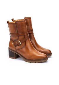 Bottines en cuir marron avec une texture lisse, un petit talon bloc, une sangle décorative et des finitions en métal doré. Équipées d'un tirette pour un enfilage facile.