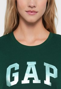 Zelené bavlněné tričko s výrazným stříbrným nápisem "GAP" vepředu, s kulatým výstřihem a měkkou texturou.