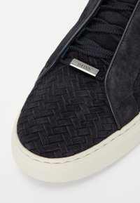 Mustat nubuk-tossut, joissa on kudottu kuvio kärjessä. Varustettu yhteensopivilla nauhoilla ja hopeisella merkkitagilla. Kermanvärinen kumipohja.