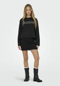 Mănușă neagră croppată cu textul "IMAGINE", asociată cu o fustă mini din denim negru. Modelul poartă cizme negre până la genunchi cu accente.
