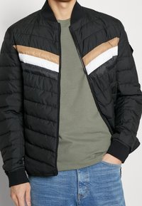 Černá péřová bunda s hnědými, bílými a černými pruhy ve tvaru chevronu na hrudi. Má žebrované manžety a zapínání na zip vpředu.