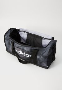 Čierna a sivá Adidas športová taška s otvoreným hlavným oddelením, ktoré zobrazuje svetlosivý interiér a čiernu úložnú kapsu, s nosnými rúčkami po stranách.