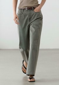 Personne portant un pantalon gris ample, un haut côtelé beige, une ceinture marron cloutée et des sandales noires, marchant sur un sol lisse dans un éclairage neutre.