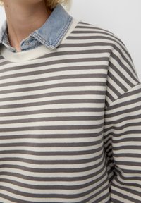 Personne portant un pull rayé gris et blanc par-dessus une chemise en denim bleu clair avec col, sur un fond blanc uni.
