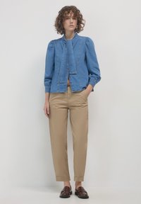 Giacca in denim con maniche a sbuffo, chiusura a bottoni e dettaglio di sciarpa annodata, abbinata a pantaloni beige e mocassini marroni.