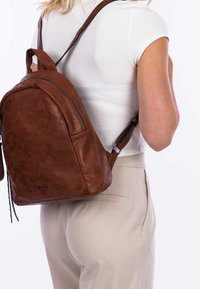 Brązowy skórzany plecak o zaokrąglonym kształcie, z zamykaną na zamek boczną kieszenią i regulowanymi paskami, noszony na białej koszuli, z teksturowanym wykończeniem.