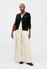 Μαύρη ζακέτα με κουμπιά και μπροστινές τσέπες, σε συνδυασμό με κρεμ παντελόνες φαρδιάς γραμμής και ψηλή μέση. Διακοσμημένη με μικρό καρφίτσα.