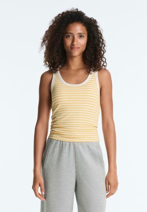 Vrouw met krullend haar, gekleed in een mouwloos geel-wit gestreept topje en grijze broek, staand tegen een effen achtergrond.