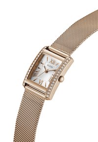 Ορθογώνιο ρολόι σε χρυσή απόχρωση με λευκό υφασμάτινο καντράν, ρωμαϊκούς αριθμούς, κρυσταλλικές λεπτομέρειες και πλεκτό λουράκι. Διαθέτει κορόνα στο πλάι.