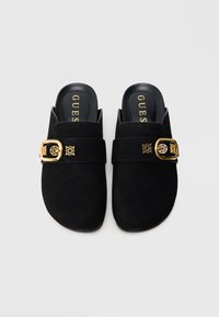 Melni zamšādas slip-on stila apavi ar platu siksnu un zelta sprādzi. Iekšējā zolīte ir melna ar iespiestu zīmola nosaukumu "GUES". Plakanas zolītes dizains.